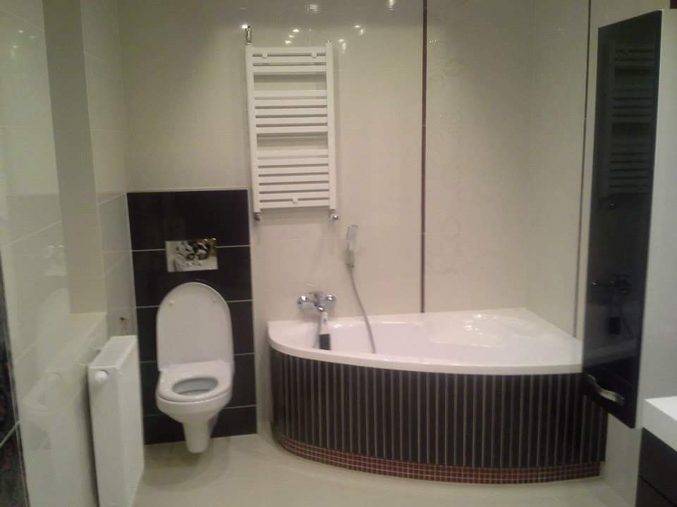 Wykończona łazienka z narożną wanną obudowaną pionowymi, brązowo-czarnymi paskami, wiszącą grzejnikiem drabinkowym oraz toaletą z czarną ścianą w tle.