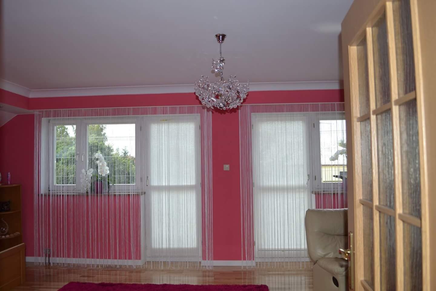 Pokój z różowymi ścianami, białymi firanami sznurkowymi, kryształowym żyrandolem i drewnianymi drzwiami z szybami.
