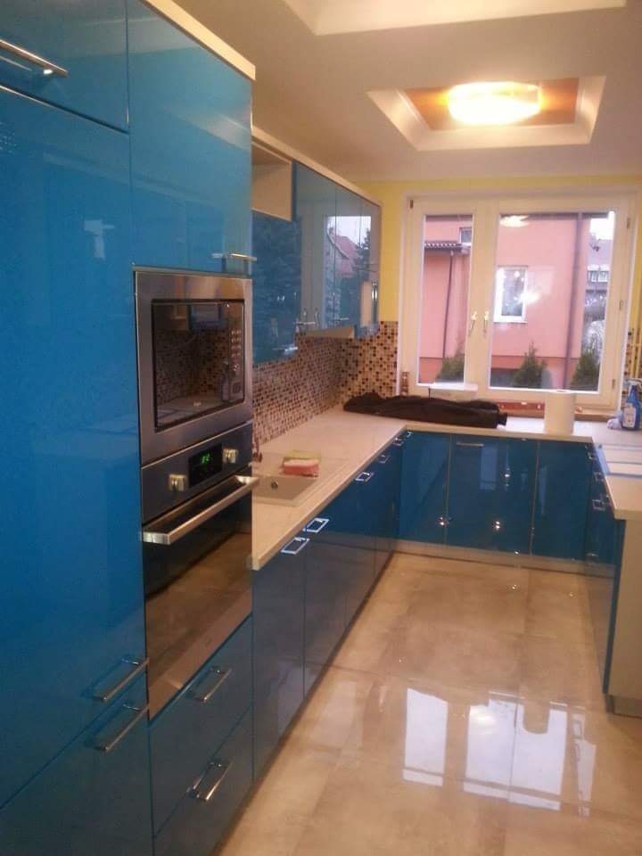 Nowoczesna kuchnia z błyszczącymi, turkusowymi szafkami, mozaiką nad blatem i jasną podłogą z płytek odbijających światło. Wbudowany piekarnik ze stali nierdzewnej. Widok na dom przez okno.