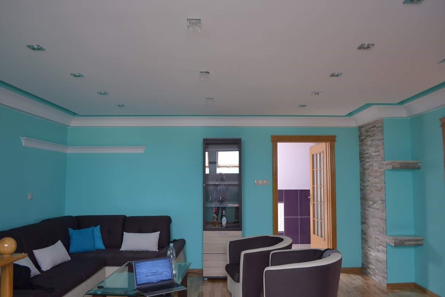 Salon w Warszawie po remoncie, z turkusowymi ścianami, białym sufitem z oświetleniem punktowym, szarą kanapą narożną, szklanym stolikiem kawowym z laptopem i dwoma fotelami.