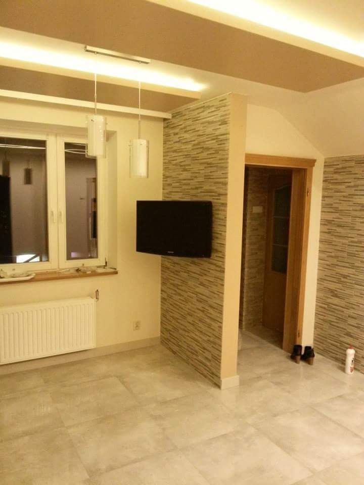 Jasne wnętrze po renowacji: płytki podłogowe imitujące kamień, ściana z dekoracyjną okładziną, telewizor na ścianie działowej, grzejnik pod oknem i wiszące lampy.