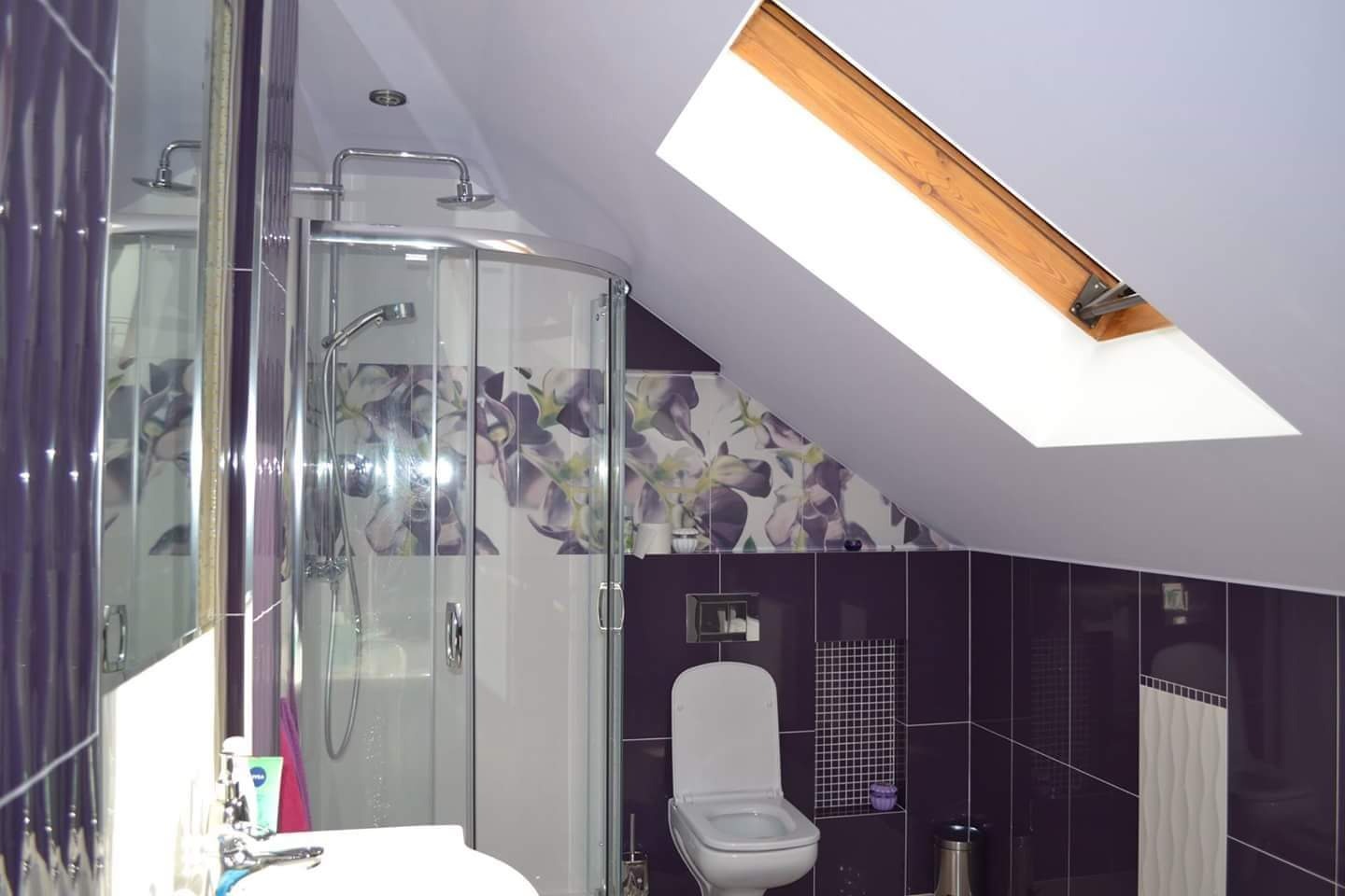 Fioletowa łazienka na poddaszu ze skośnym oknem dachowym, kabiną prysznicową z kwiatowym motywem i toaletą.