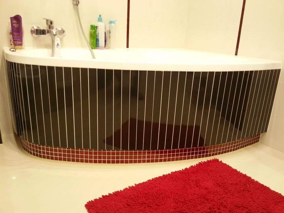 Półokrągła wanna obudowana czarnymi płytkami z białymi fugami, wykończona mozaiką w odcieniach czerwieni, z baterią i kosmetykami na krawędzi oraz czerwonym dywanikiem na podłodze.
