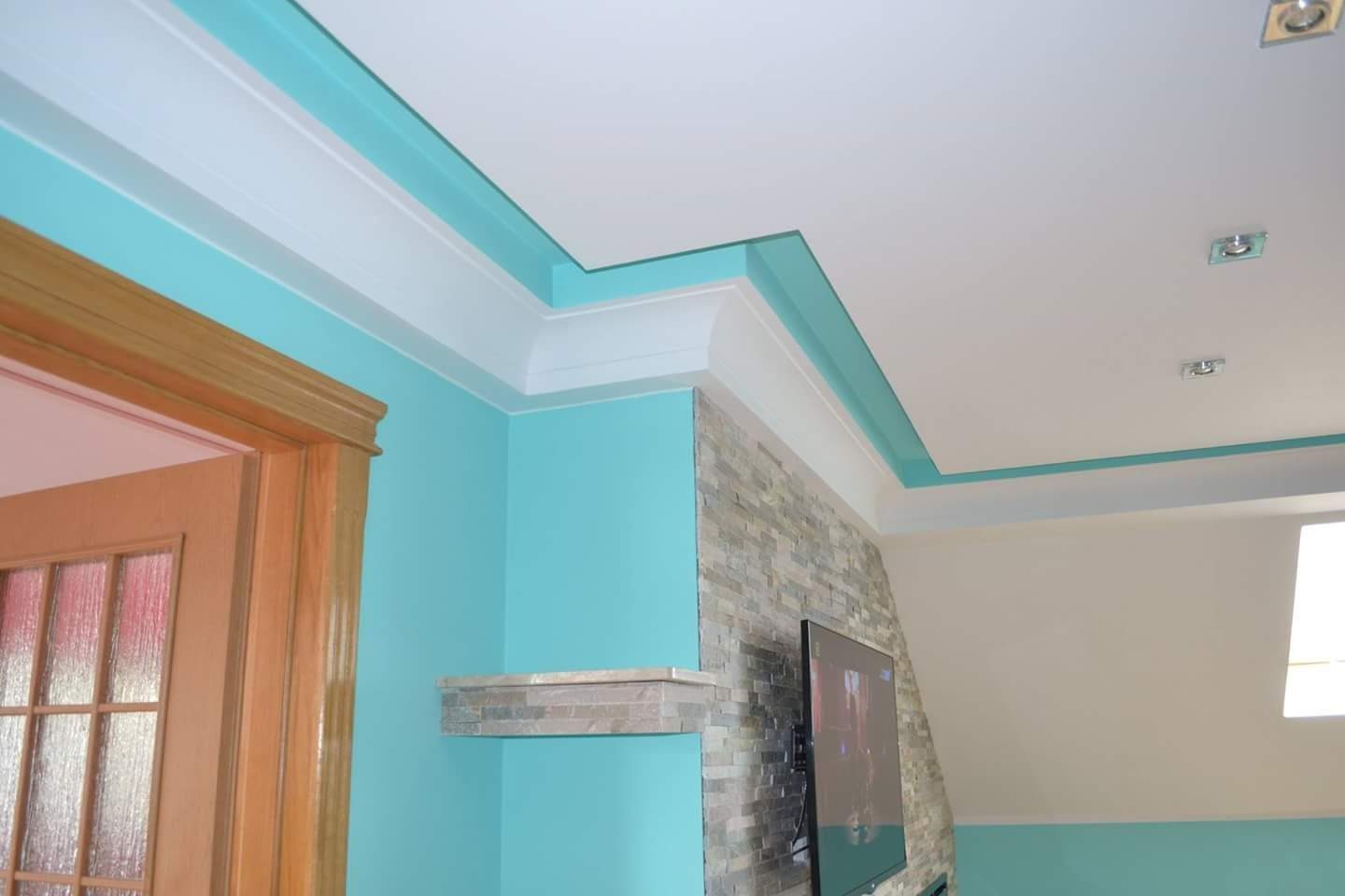 Turkusowa ściana z białym sztukaterią sufitową, kamienna ściana z telewizorem oraz drewniane drzwi z szybami w wyremontowanym pokoju.