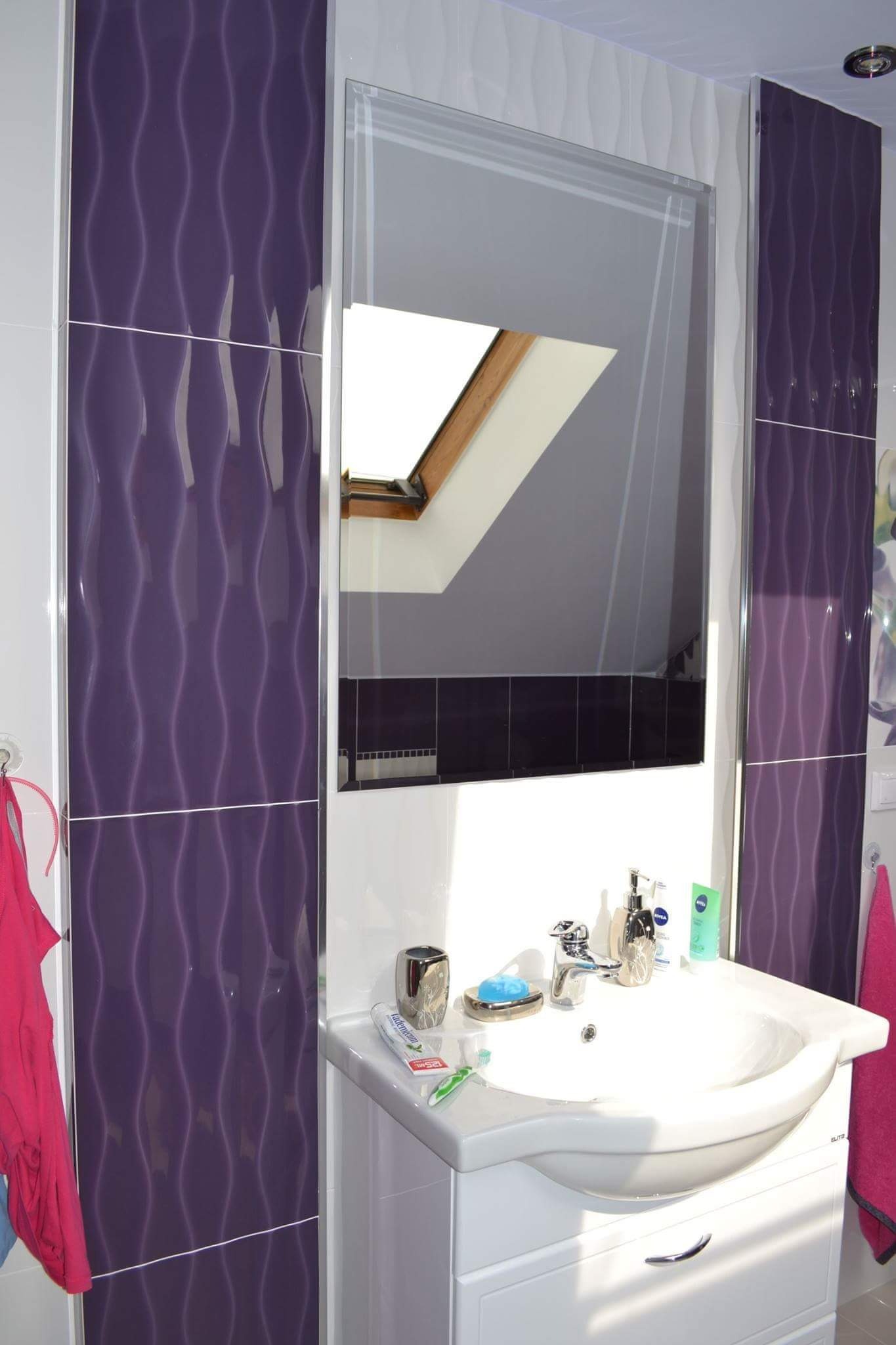 Biała umywalka z szafką w łazience z fioletowymi, falistymi płytkami, odbicie okna dachowego w lustrze.