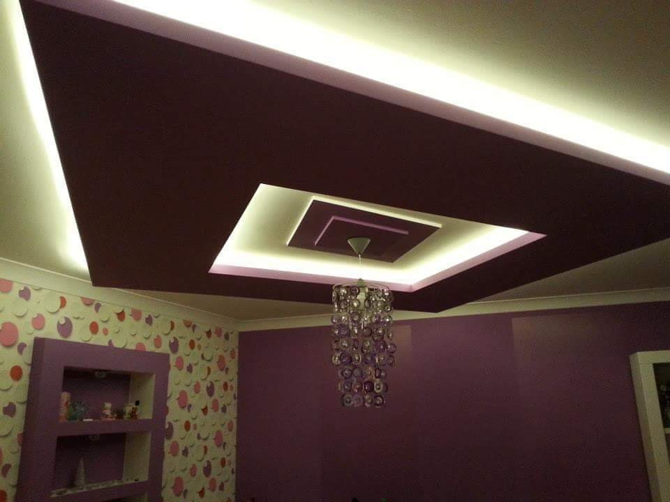 Wielopoziomowy, podwieszany sufit w odcieniach fioletu z oświetleniem LED, uzupełniony dekoracyjnym żyrandolem i wzorzystą tapetą na ścianie.