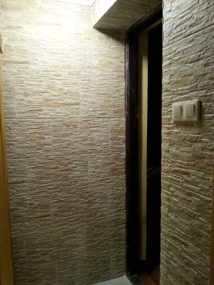 Wąski korytarz wykończony panelami ściennymi imitującymi kamień, z widocznym włącznikiem światła i ościeżnicą drzwi.