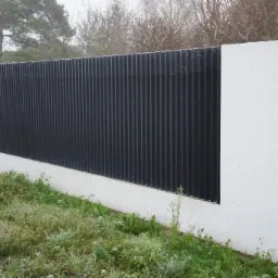 Nowoczesne ogrodzenie z pionowych, czarnych paneli umieszczonych pomiędzy białymi, betonowymi słupkami, otoczone zielenią.