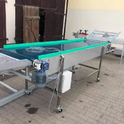 Stalowa linia produkcyjna z zielonymi elementami transportującymi, wyposażona w silnik elektryczny i kółka, ustawiona na zewnątrz budynku z drewnianymi drzwiami.