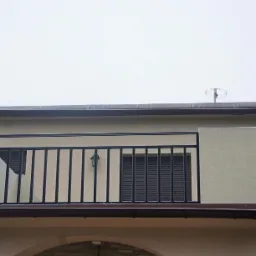 Balkon z czarną, metalową balustradą o prostym wzorze, widoczny z dołu na tle szarego nieba, z anteną na dachu budynku.