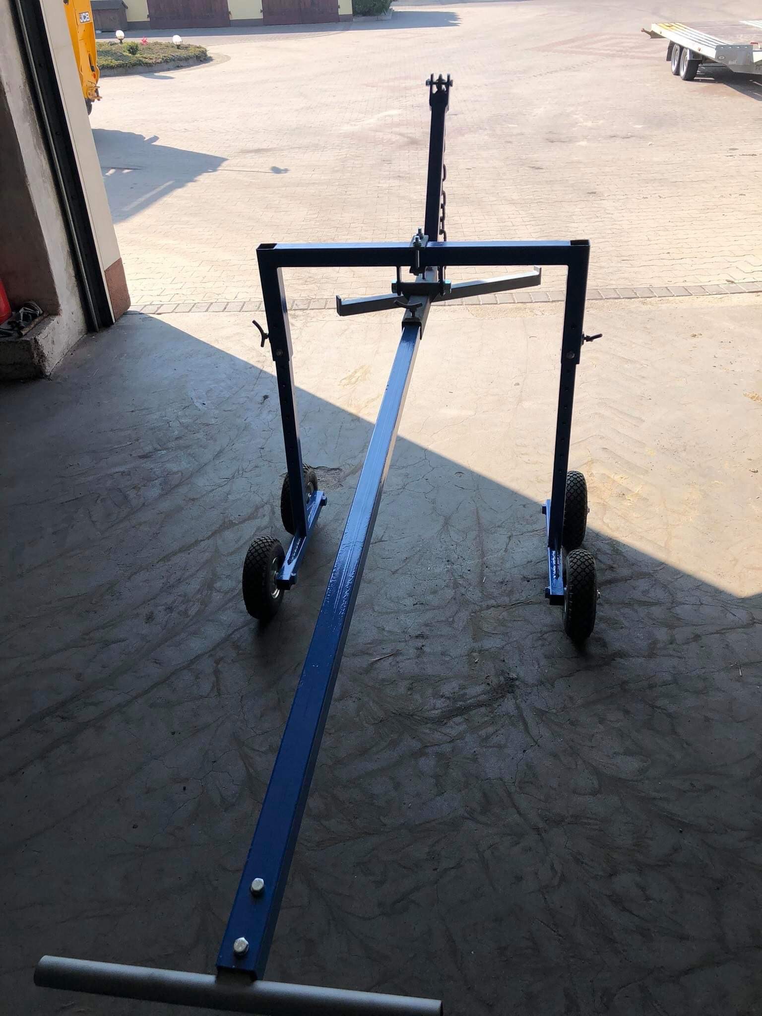 Wózek warsztatowy do montażu i transportu elementów stalowych, pomalowany na niebiesko, z kołami terenowymi, stojący w warsztacie z betonową podłogą. W tle widoczny budynek i przyczepa.