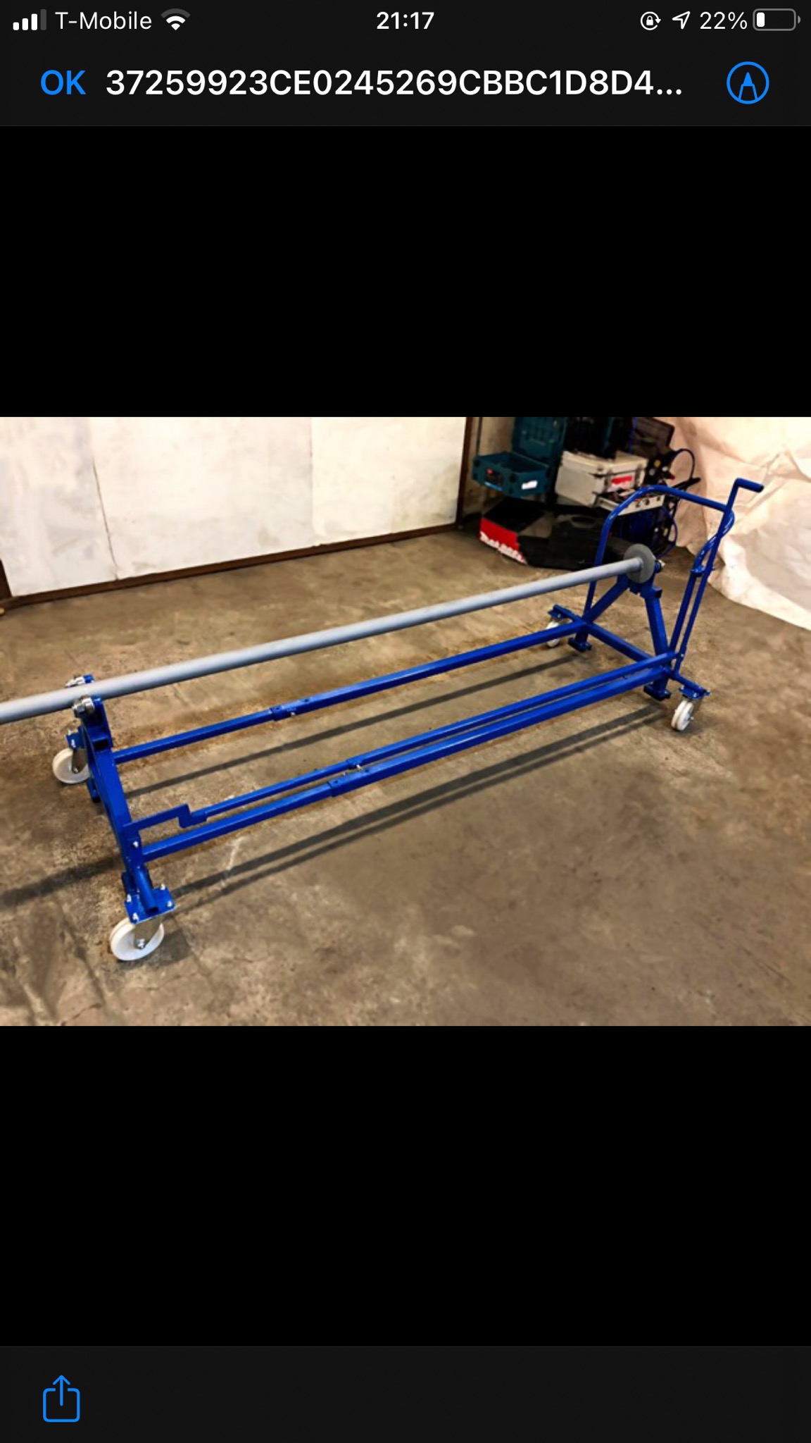 Niebieski, metalowy stojak na kółkach z zamontowaną szarą rurą, widoczny na tle betonowej podłogi i białej ściany, z skrzynkami narzędziowymi w tle.