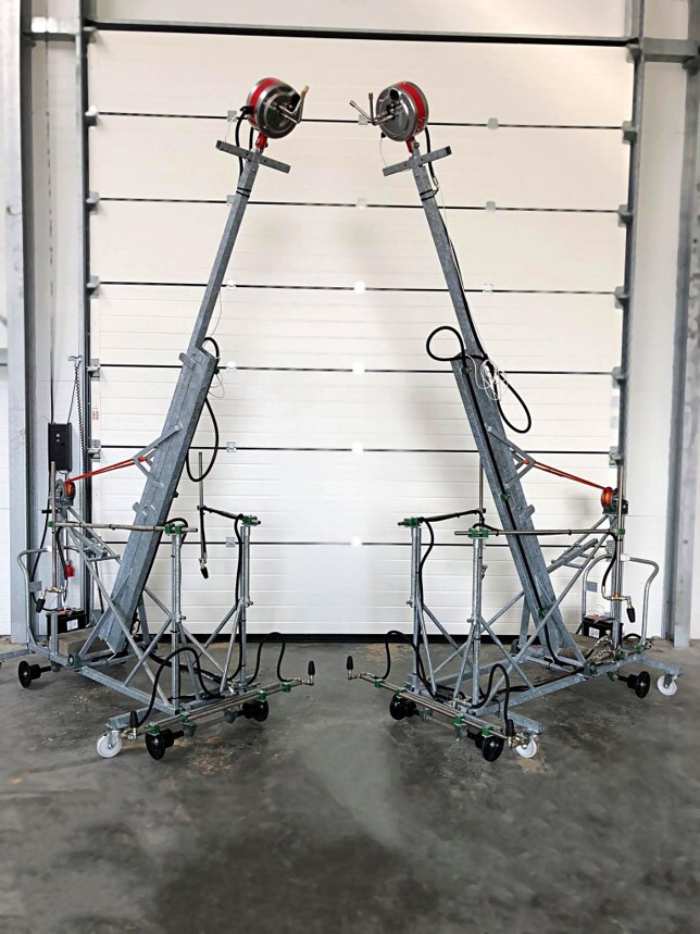 Dwa identyczne, mobilne stanowiska spawalnicze z podajnikami drutu, ustawione naprzeciwko zamkniętej, segmentowej bramy garażowej.