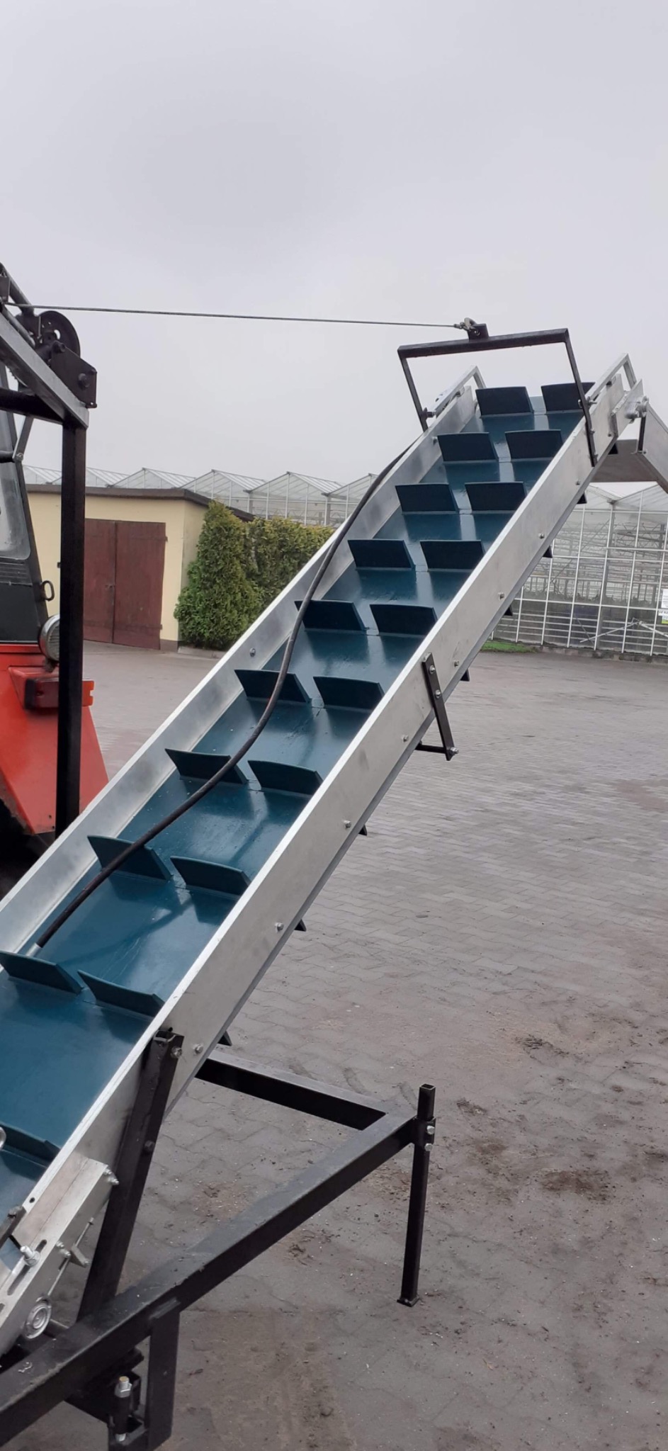 Podnośnik taśmowy z niebieską taśmą i aluminiową konstrukcją, ustawiony pod kątem, z widocznym fragmentem czerwonego ciągnika i szklarnią w tle.