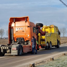 RATOWNICTWO DROGOWE ADMAR S.C - Pomarańczowy ciągnik siodłowy MAN holowany przez żółtą lawetę na poboczu drogi poza miastem, widoczny mężczyzna w kurtce i czapce przy pojeździe.