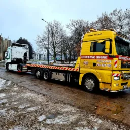 Żółta laweta firmy ADMAR z załadowaną białą kabiną ciężarówki Renault na mokrej, zimowej drodze w mieście.