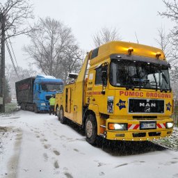RATOWNICTWO DROGOWE ADMAR S.C - Żółta ciężarówka pomocy drogowej MAN z napisem 'POMOC DROGOWA' holuje niebieską ciężarówkę w zimowej scenerii, widoczne ślady opon i stóp na śniegu.