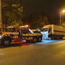 RATOWNICTWO DROGOWE ADMAR S.C - Ciężarówka holownicza z włączonymi światłami ostrzegawczymi holuje białą ciężarówkę na oświetlonej ulicy miejskiej nocą.