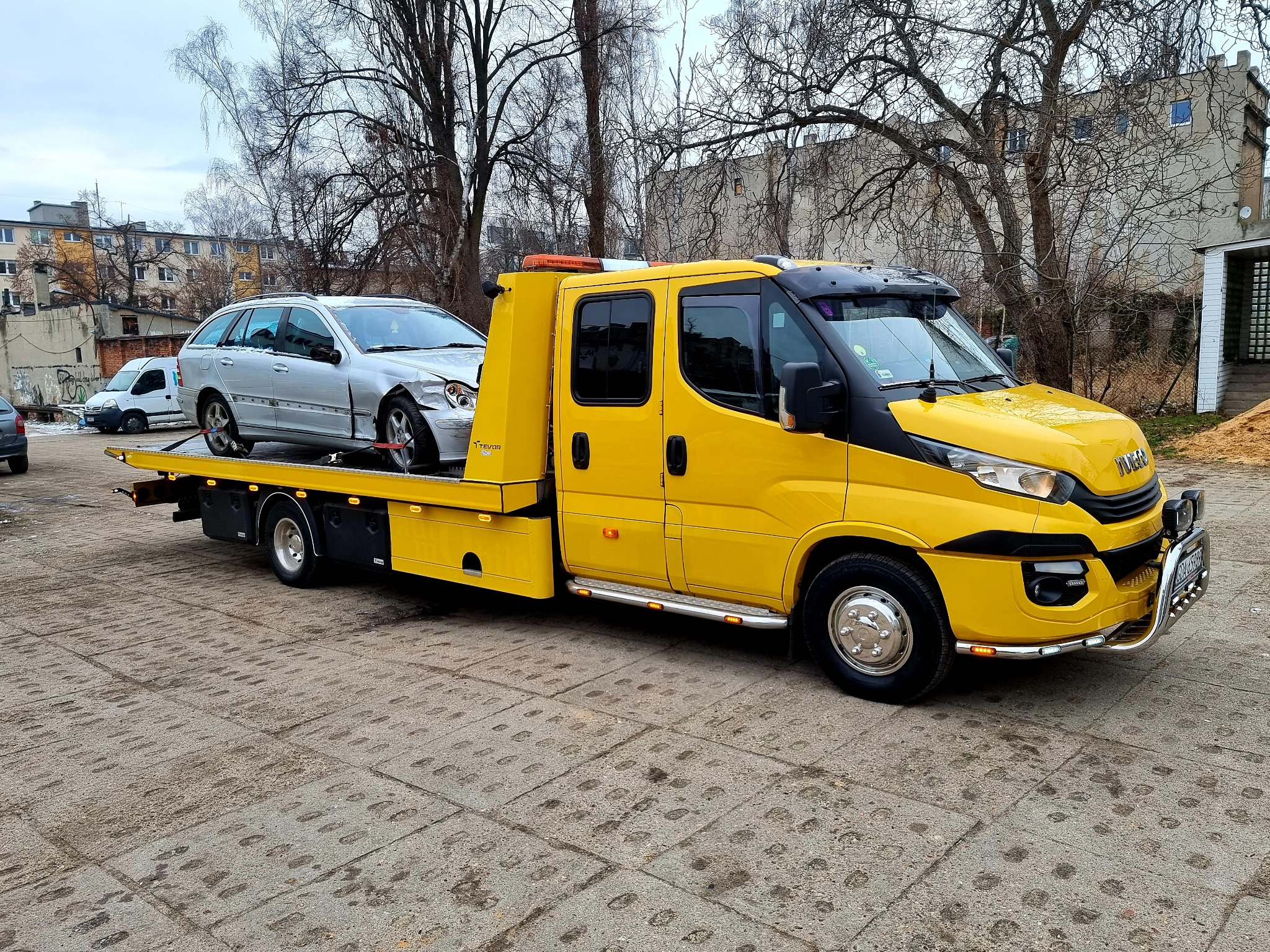 Żółta laweta Iveco z uszkodzonym srebrnym kombi Mercedes na platformie, zaparkowana na betonowym placu w pochmurny dzień.
