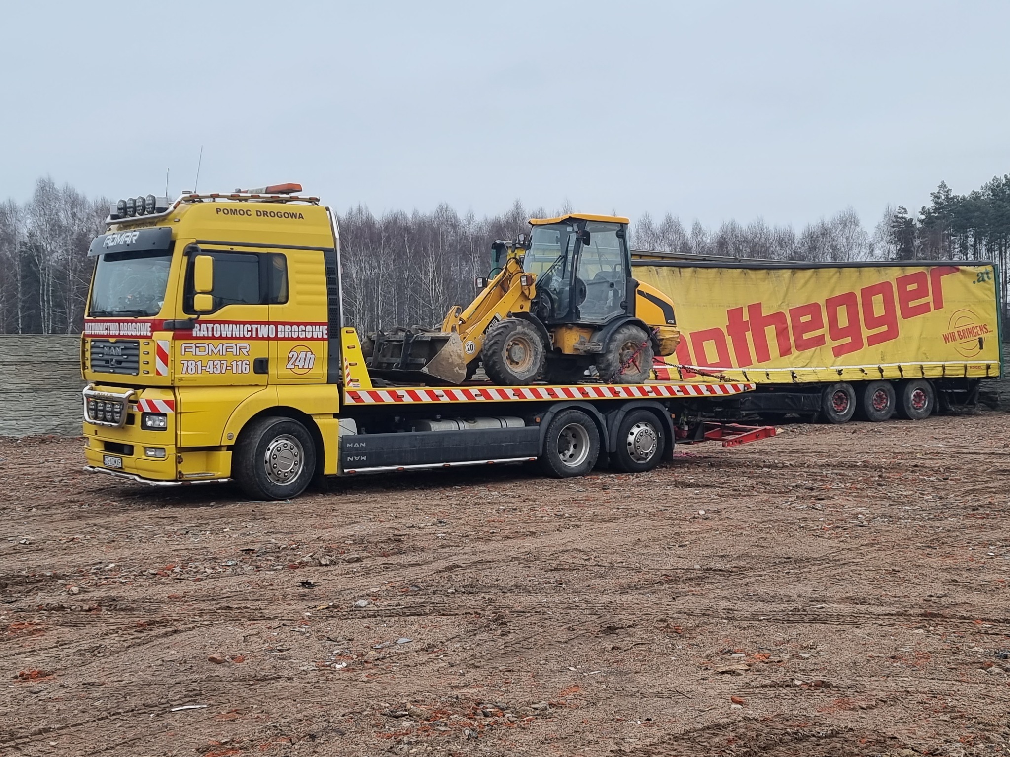 Żółta laweta z Dąbrówki Wielkiej przewozi żółtą koparko-ładowarkę na tle zalesionego terenu. Na naczepie lawety widoczna reklama firmy.