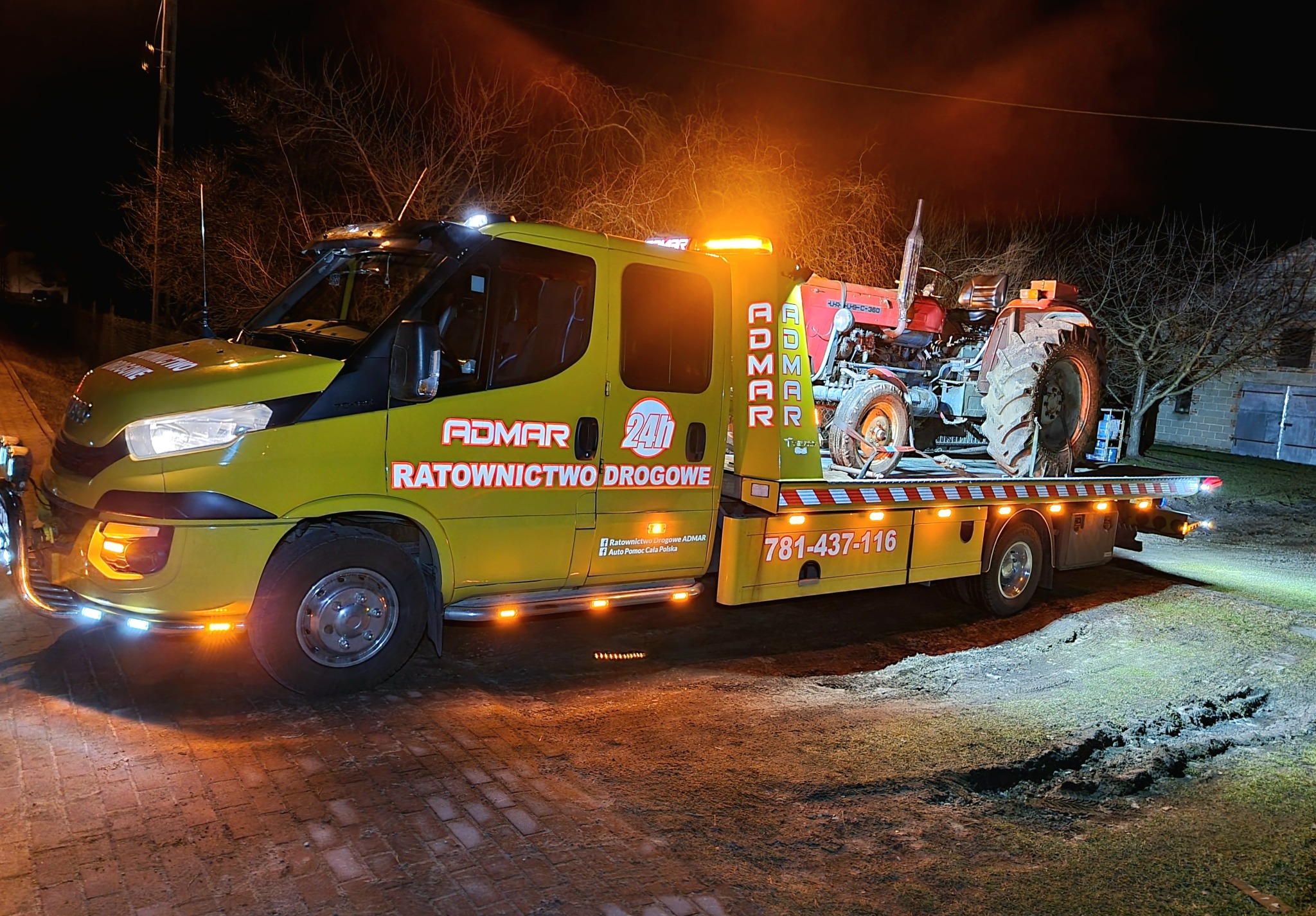 Żółta laweta z napisem 'ADMAR Ratownictwo Drogowe' przewozi czerwony traktor na platformie w nocy, z włączonymi światłami ostrzegawczymi.