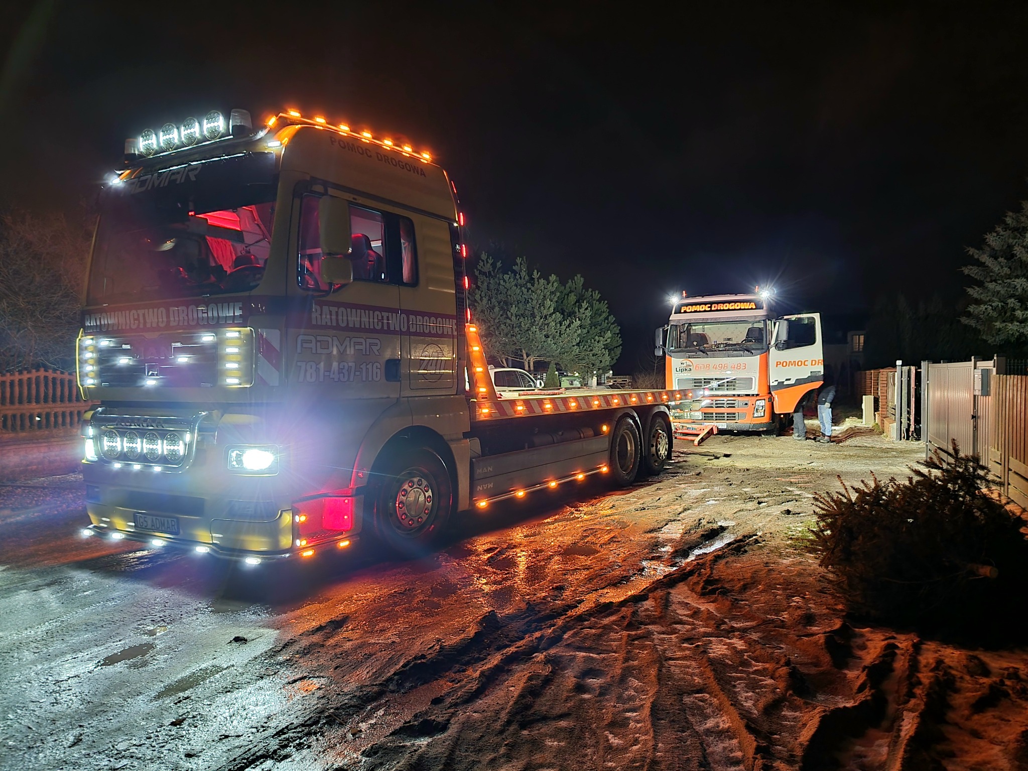 Dwie lawety pomocy drogowej w akcji nocą, oświetlone licznymi lampami, na błotnistym podłożu, z widocznym drzewem iglastym w pobliżu.