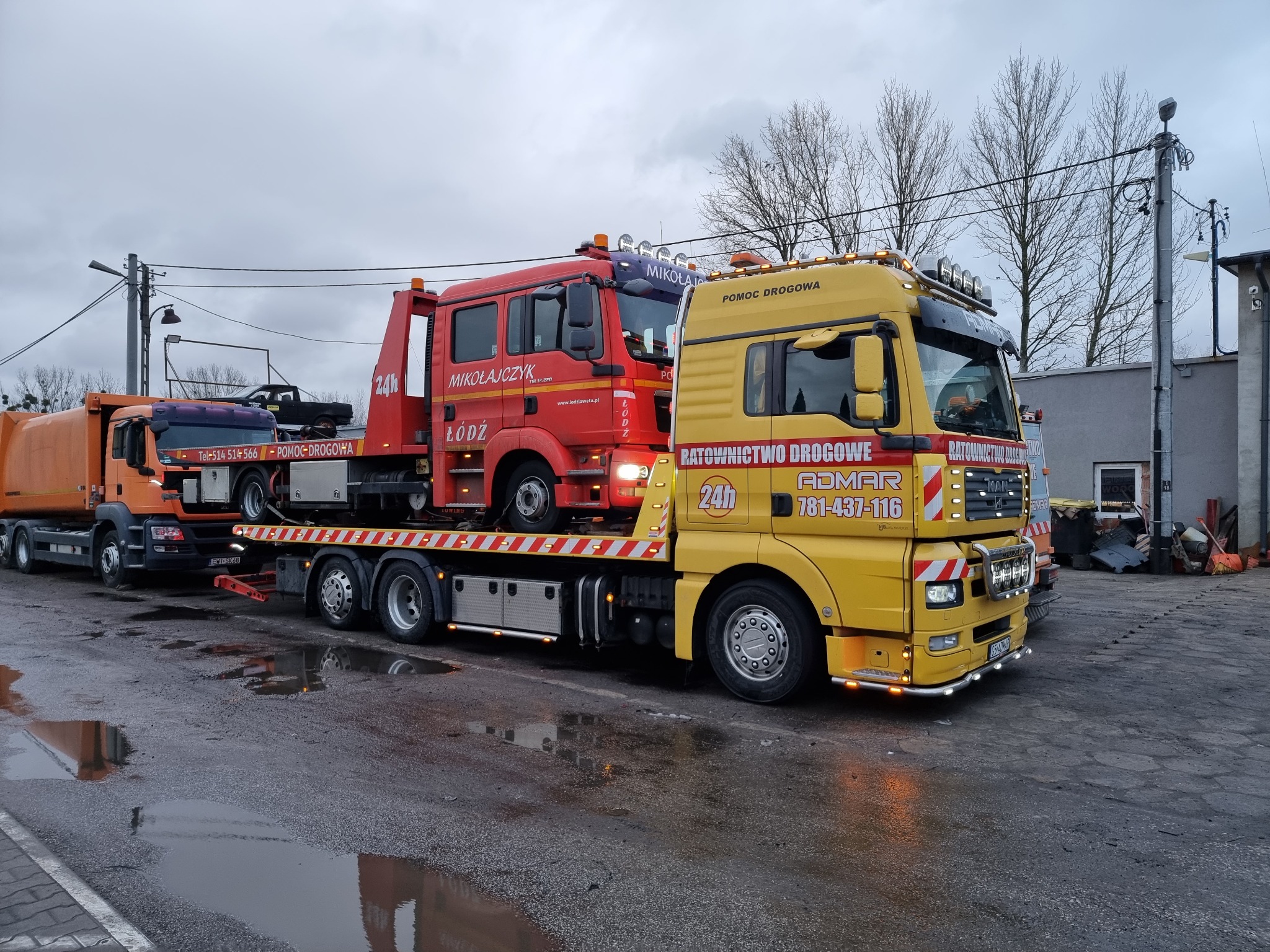 Żółta ciężarówka pomocy drogowej z czerwonym pojazdem na platformie, zaparkowana na mokrym, popękanym asfalcie w pochmurny dzień.
