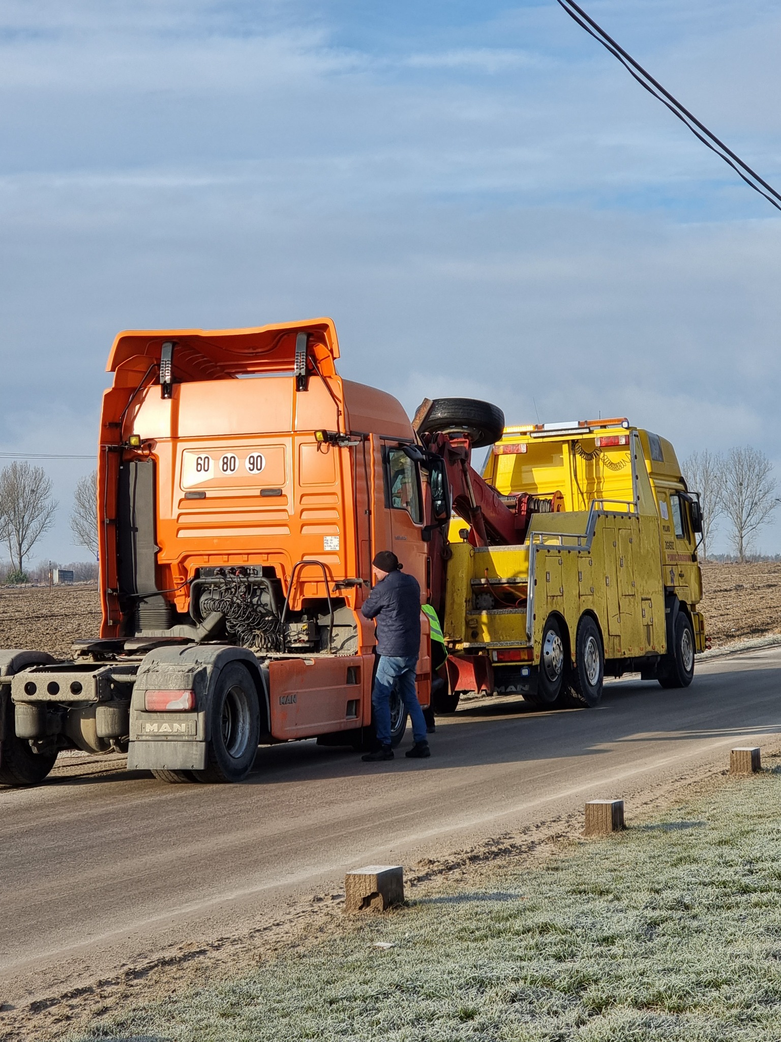 Pomarańczowy ciągnik siodłowy MAN holowany przez żółtą lawetę na poboczu drogi poza miastem, widoczny mężczyzna w kurtce i czapce przy pojeździe.