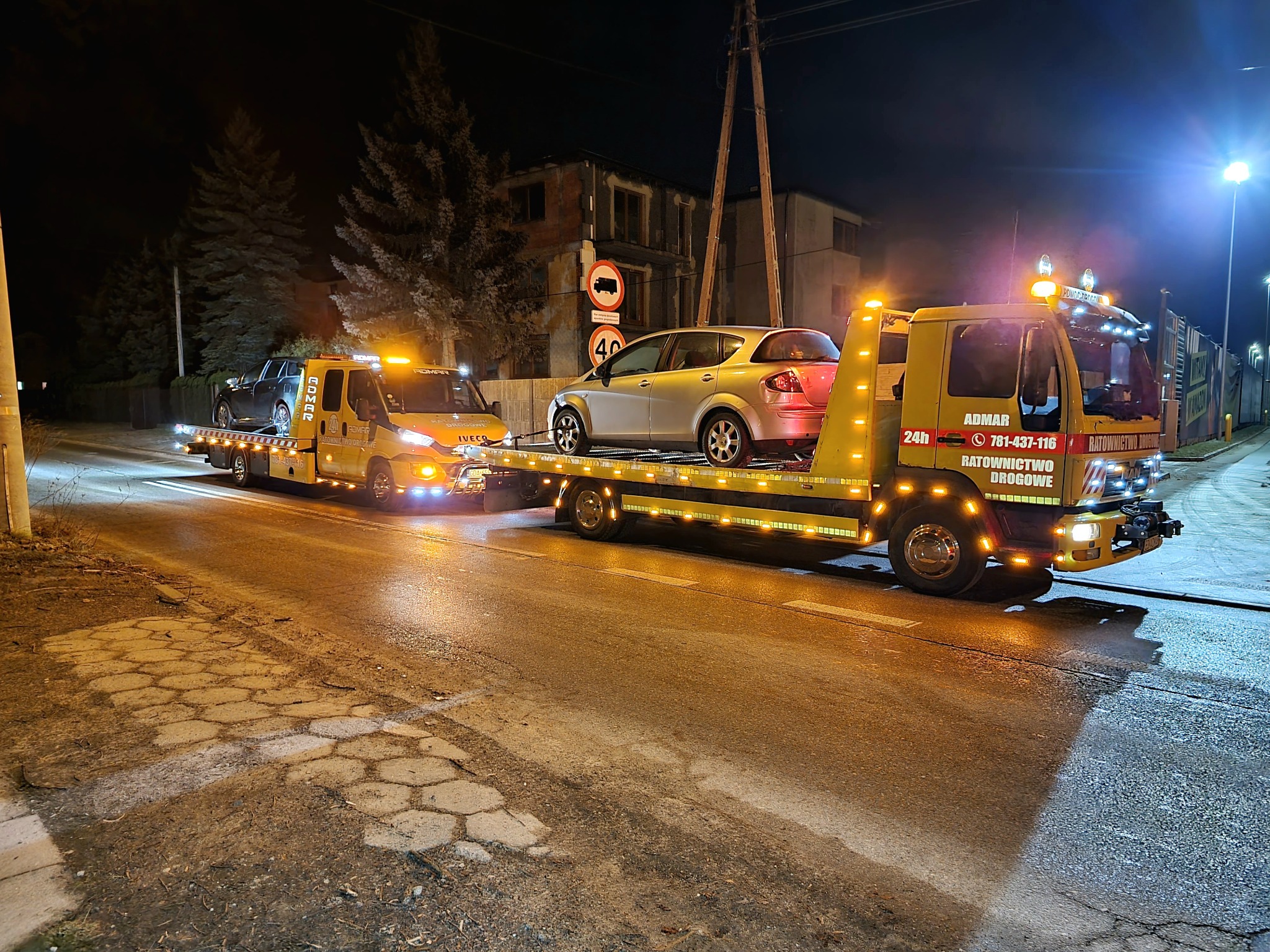 Dwie żółte lawety z włączonymi światłami ostrzegawczymi transportują samochody osobowe nocą na oświetlonej ulicy. Na drugiej lawecie srebrny samochód. W tle budynek w trakcie remontu.