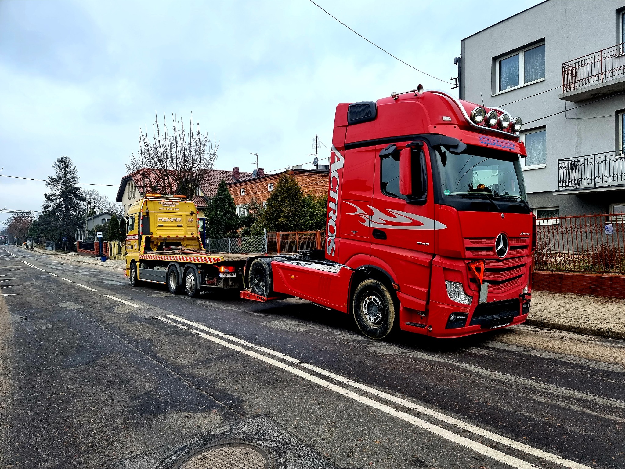 Czerwona ciężarówka marki Mercedes-Benz Actros z napisem 'Actros' na kabinie, holująca żółtą lawetę na dwupasmowej drodze w mieście, na tle szarego budynku mieszkalnego.