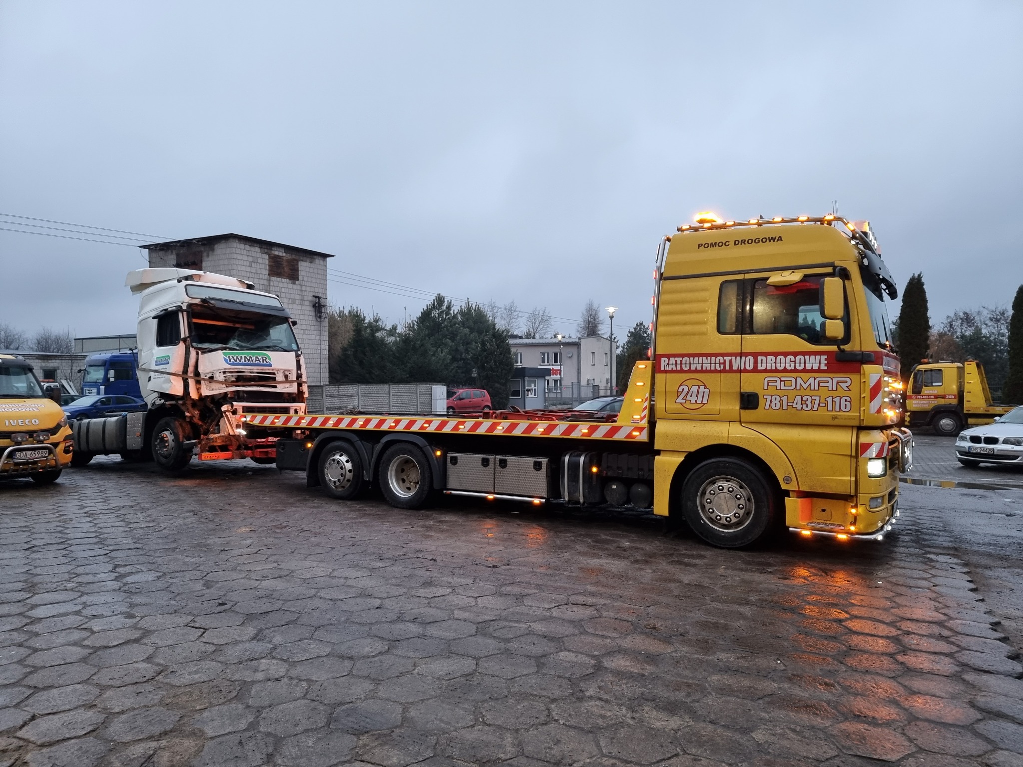 Żółta ciężarówka pomocy drogowej z napisem 'RATOWNICTWO DROGOWE ADMAR 781-437-116' holuje uszkodzoną białą ciężarówkę z widocznym logo 'IW-MAR' na mokrym brukowanym placu w pochmurny dzień.