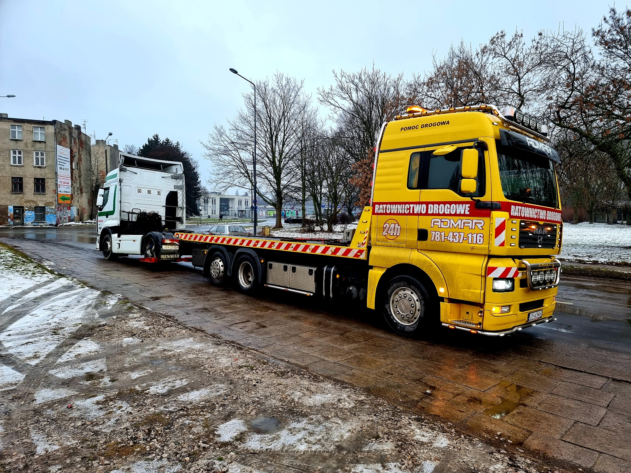Żółta laweta firmy ADMAR z załadowaną białą kabiną ciężarówki Renault na mokrej, zimowej drodze w mieście.