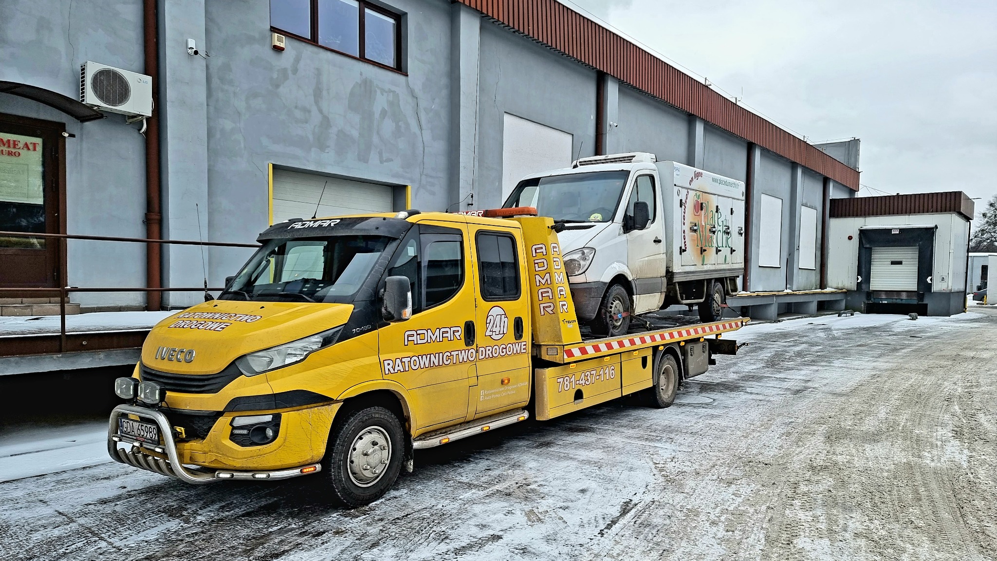 Żółta laweta firmy ADMAR z załadowanym białym busem chłodnią na tle magazynu z rampą załadunkową, zimowa sceneria.