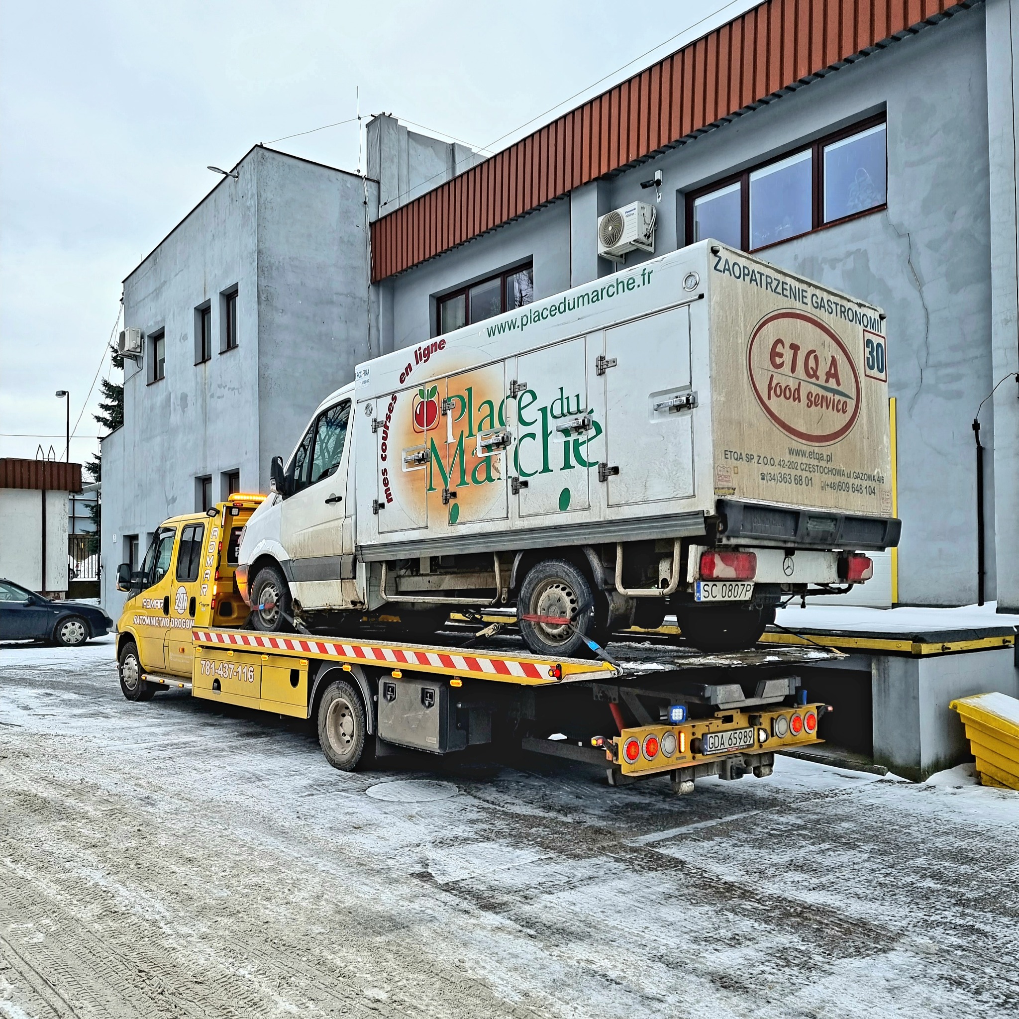 Żółta laweta z białym busem chłodnią na platformie, zaparkowana na ośnieżonym placu przed szarym budynkiem, widoczne numery rejestracyjne lawety i busa.