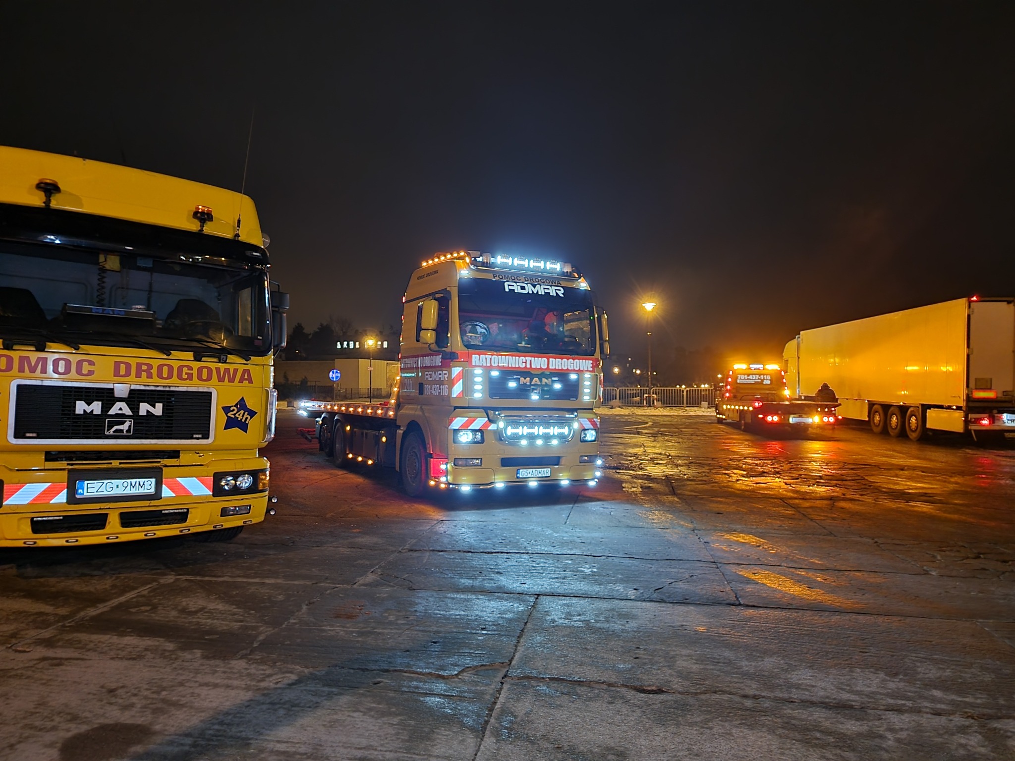 Żółte ciężarówki pomocy drogowej MAN na mokrym placu w nocy, z włączonymi światłami ostrzegawczymi i tablicami informacyjnymi.
