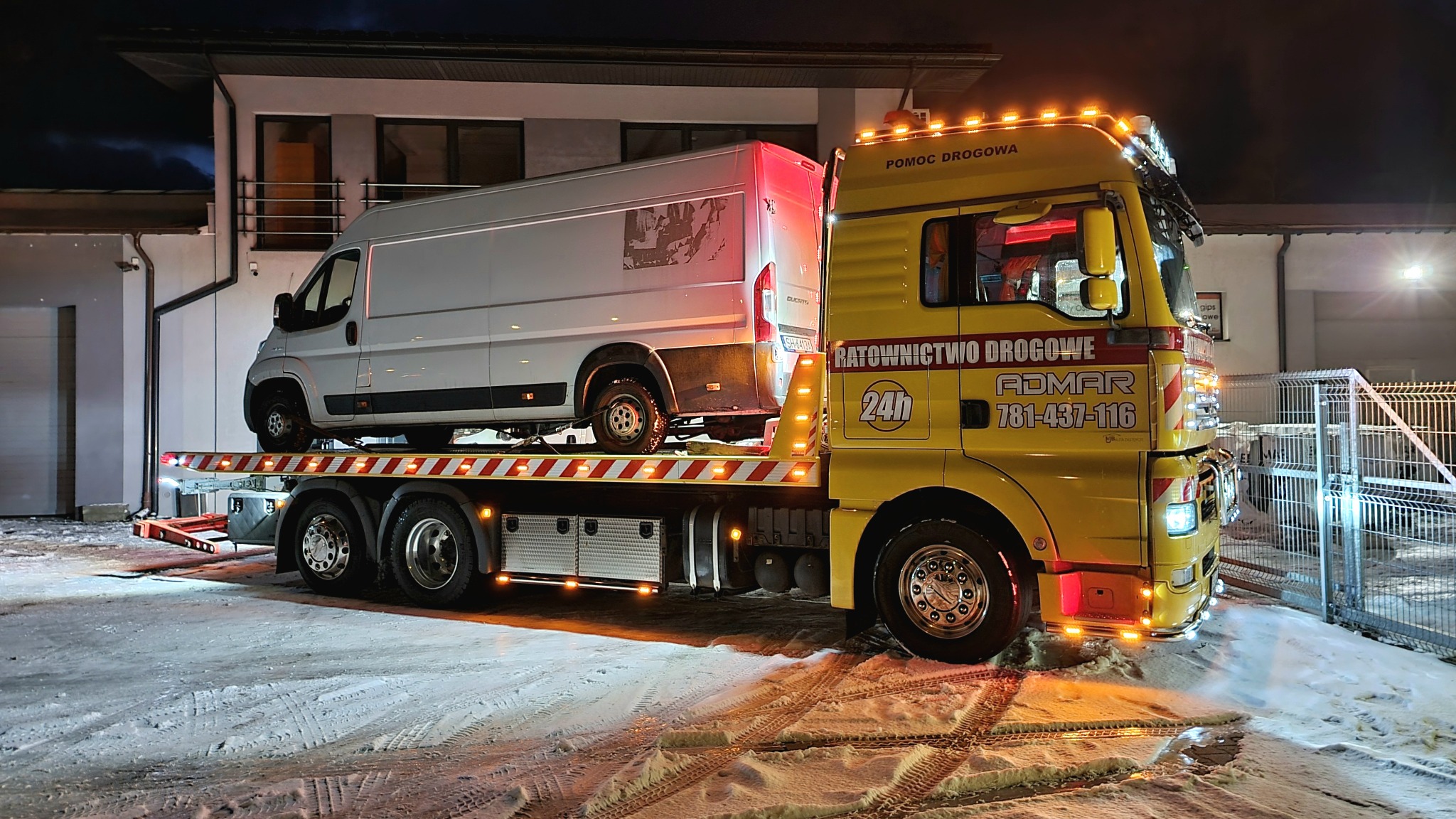 Żółta laweta z napisem 'RATOWNICTWO DROGOWE' i numerem telefonu przewozi białego busa na tle nowoczesnego domu w zimowej scenerii.