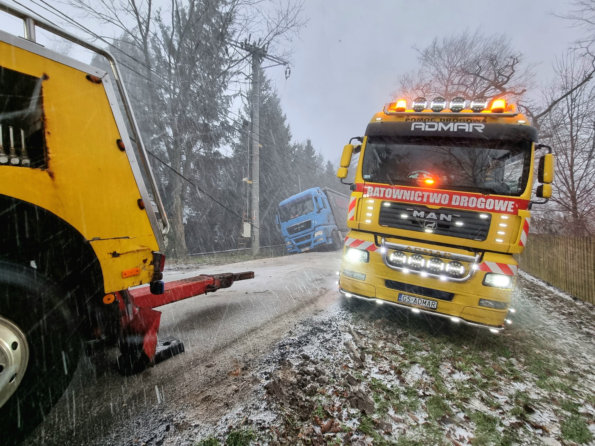 Żółta laweta z włączonymi światłami awaryjnymi na zasypanej śniegiem drodze, w tle widoczna druga laweta