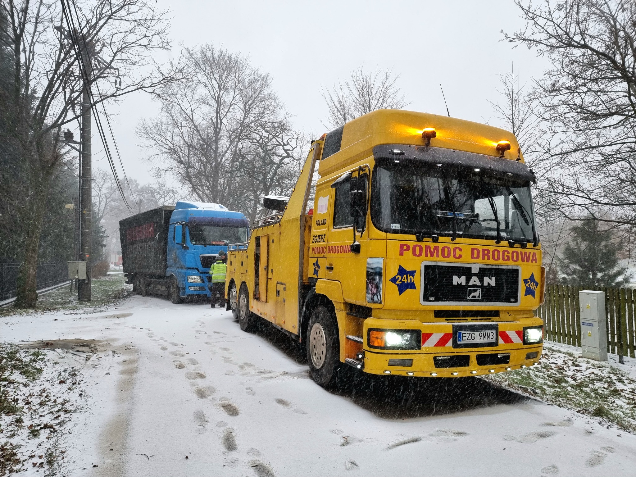 Żółta ciężarówka pomocy drogowej MAN z napisem 'POMOC DROGOWA' holuje niebieską ciężarówkę w zimowej scenerii, widoczne ślady opon i stóp na śniegu.