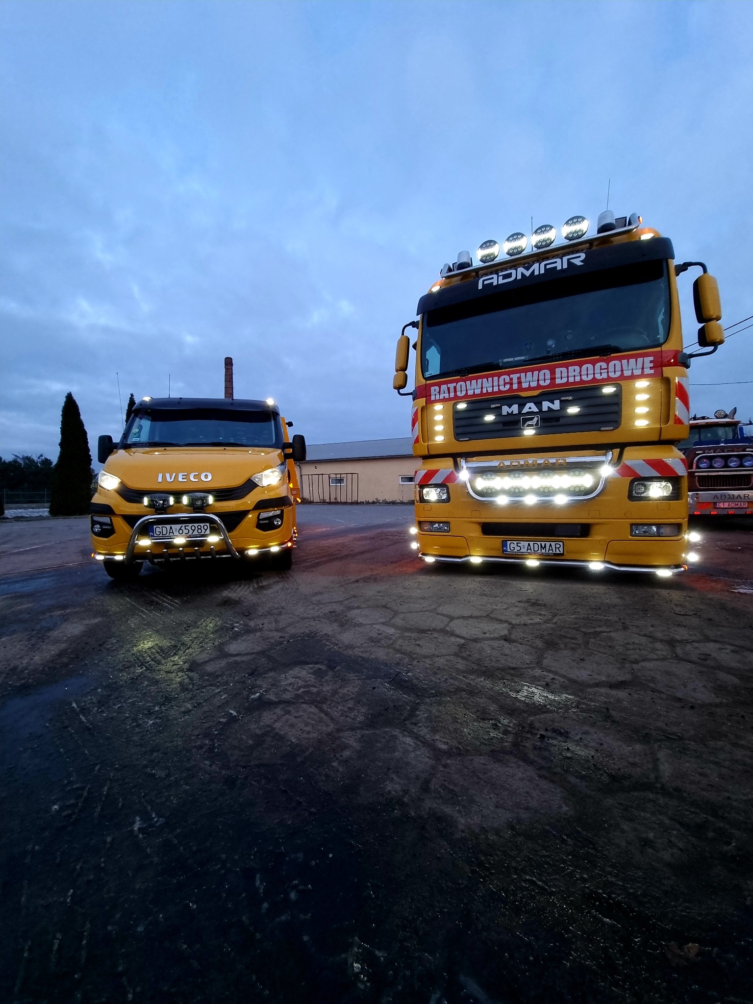 Dwa żółte pojazdy pomocy drogowej firmy Admar, Iveco i MAN, stojące na placu o zmierzchu, z włączonymi światłami i widocznym kominem w tle.