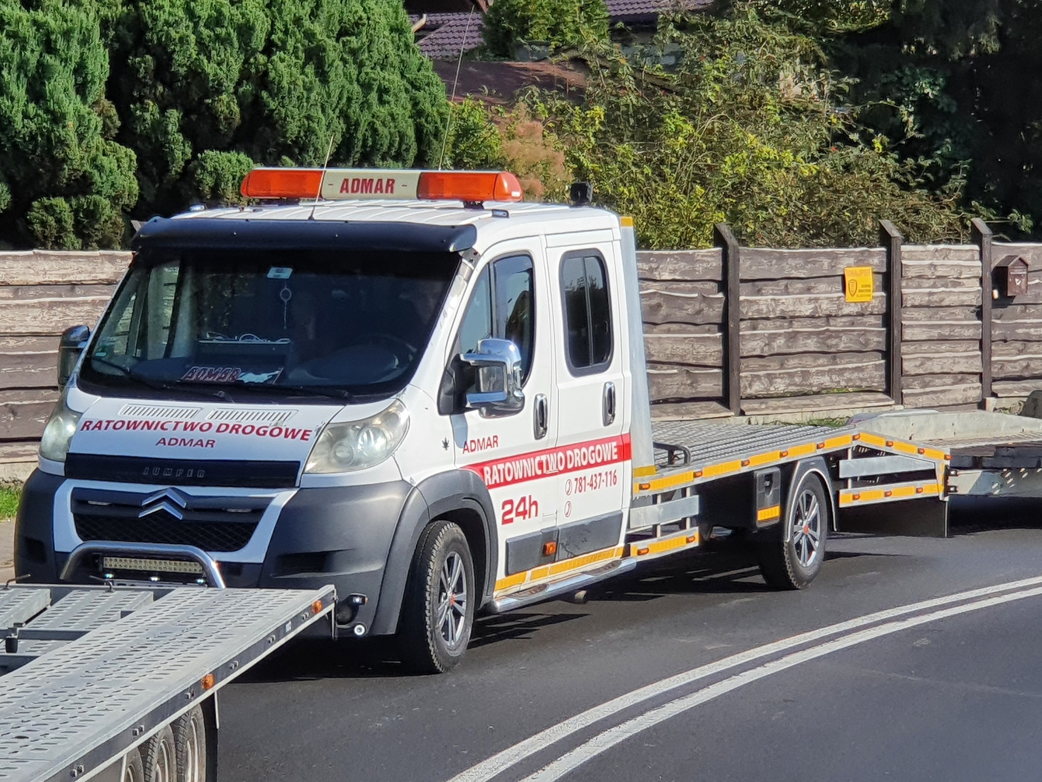 Biała laweta z pomarańczowymi światłami ostrzegawczymi na dachu, zaparkowana na ulicy w słoneczny dzień, z widocznym fragmentem platformy załadunkowej i logo firmy 'ADMAR'