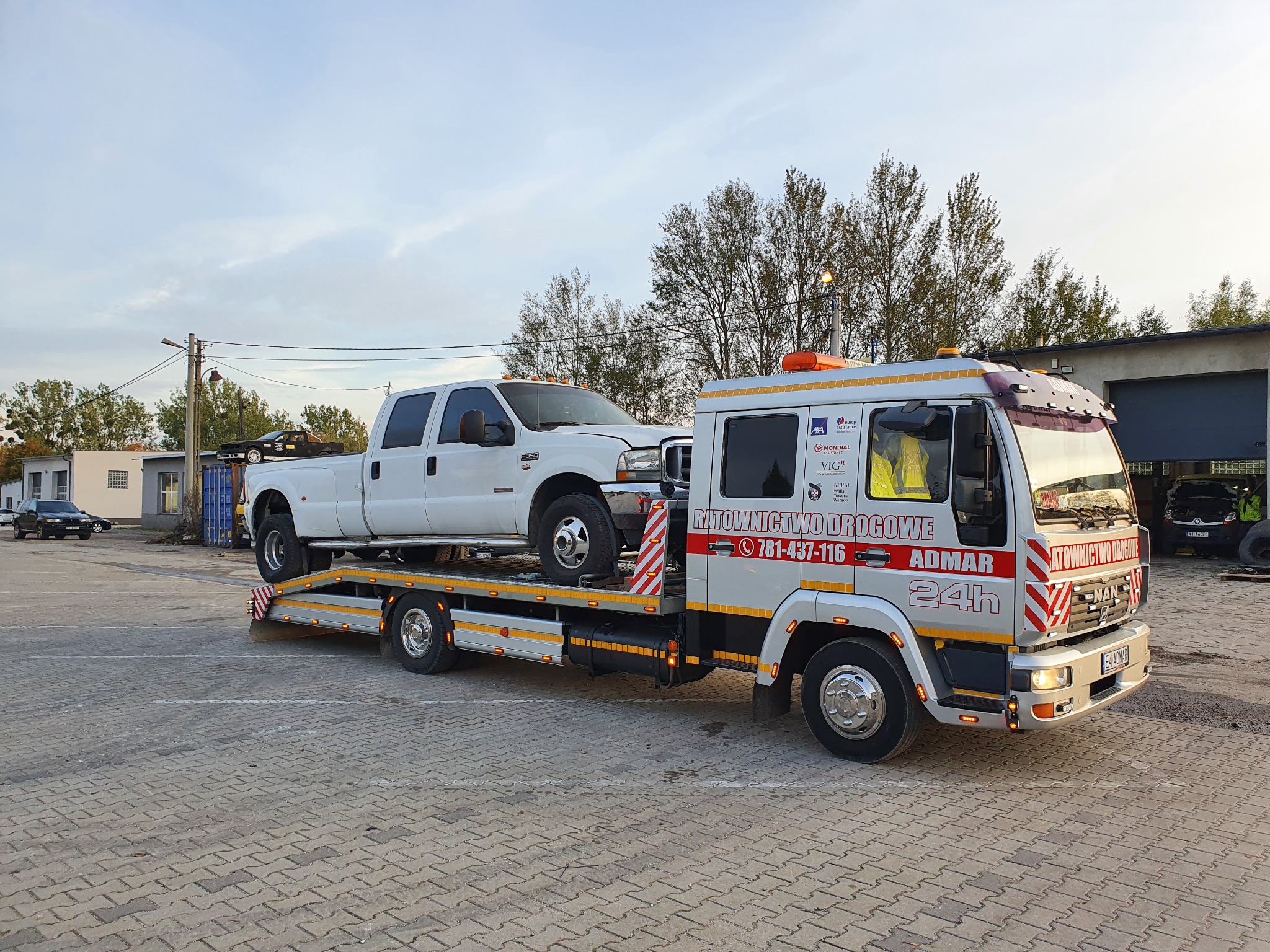 Biały pickup Ford F-350 na platformie lawety ADMAR z napisem 'Ratownictwo Drogowe 24h' na tle kostki brukowej i budynków.