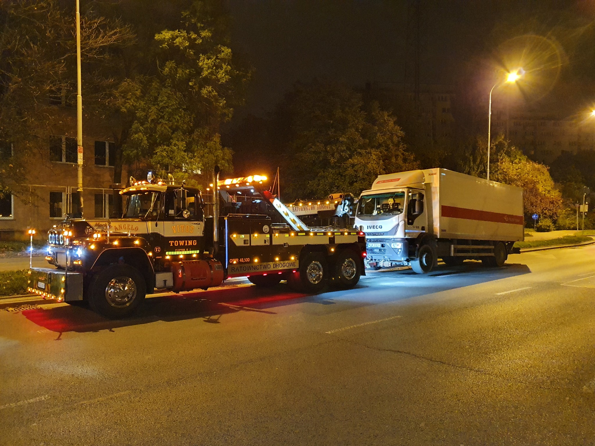 Ciężarówka holownicza z włączonymi światłami ostrzegawczymi holuje białą ciężarówkę na oświetlonej ulicy miejskiej nocą.