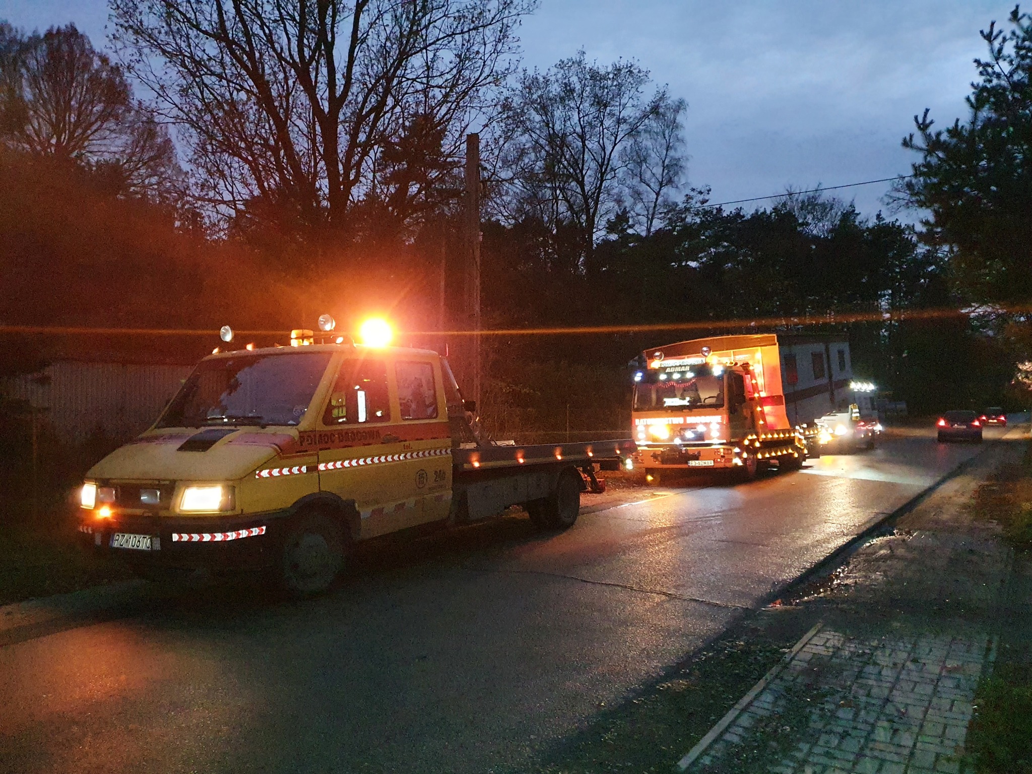 Żółta laweta z włączonymi światłami ostrzegawczymi holuje ciężarówkę pomocy drogowej na mokrej, oświetlonej ulicy o zmierzchu.