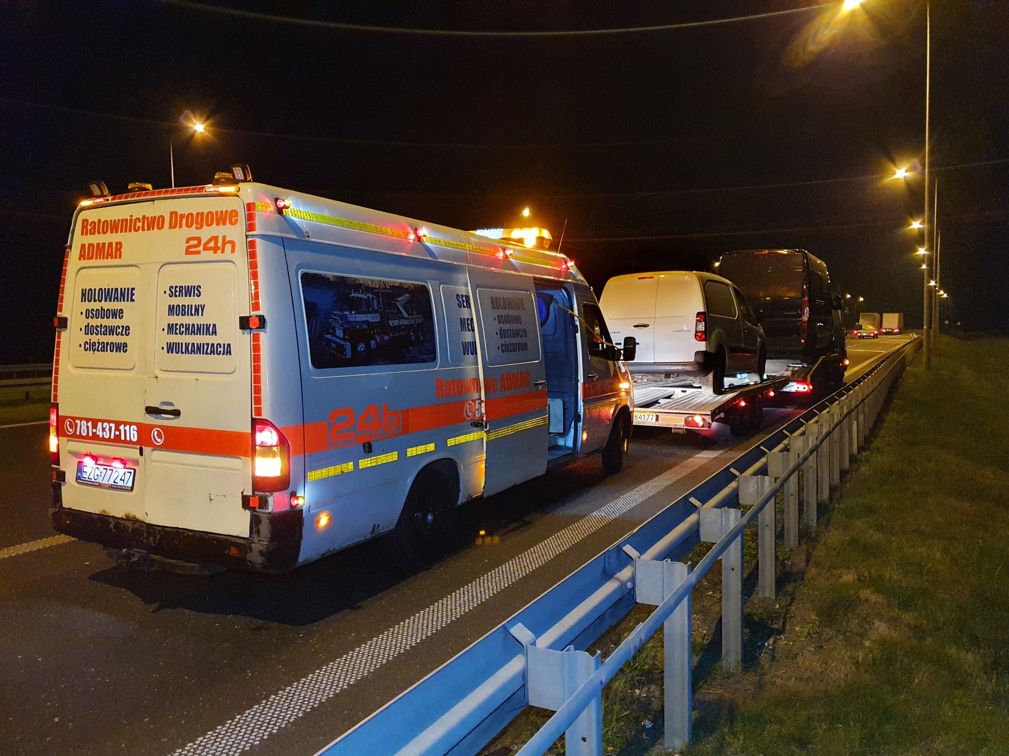 Laweta z otwartymi drzwiami na autostradzie, przewożąca dwa busy na przyczepie w nocy, oświetlona światłami ulicznymi, z widocznym napisem 'Ratownictwo Drogowe ADMAR 24h' na pojeździe.