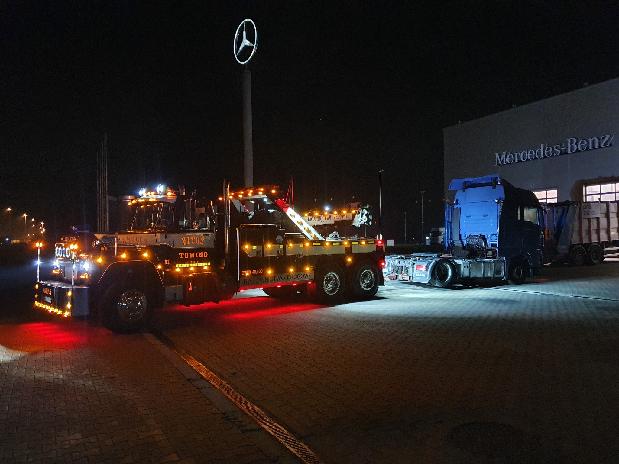 Ciężka laweta z napisem 'Ratownictwo Drogowe' i 'Vitos Towing' oświetlona czerwonymi światłami, stojąca nocą na brukowanym placu przed budynkiem Mercedes-Benz, z widocznym logo marki na słupie.