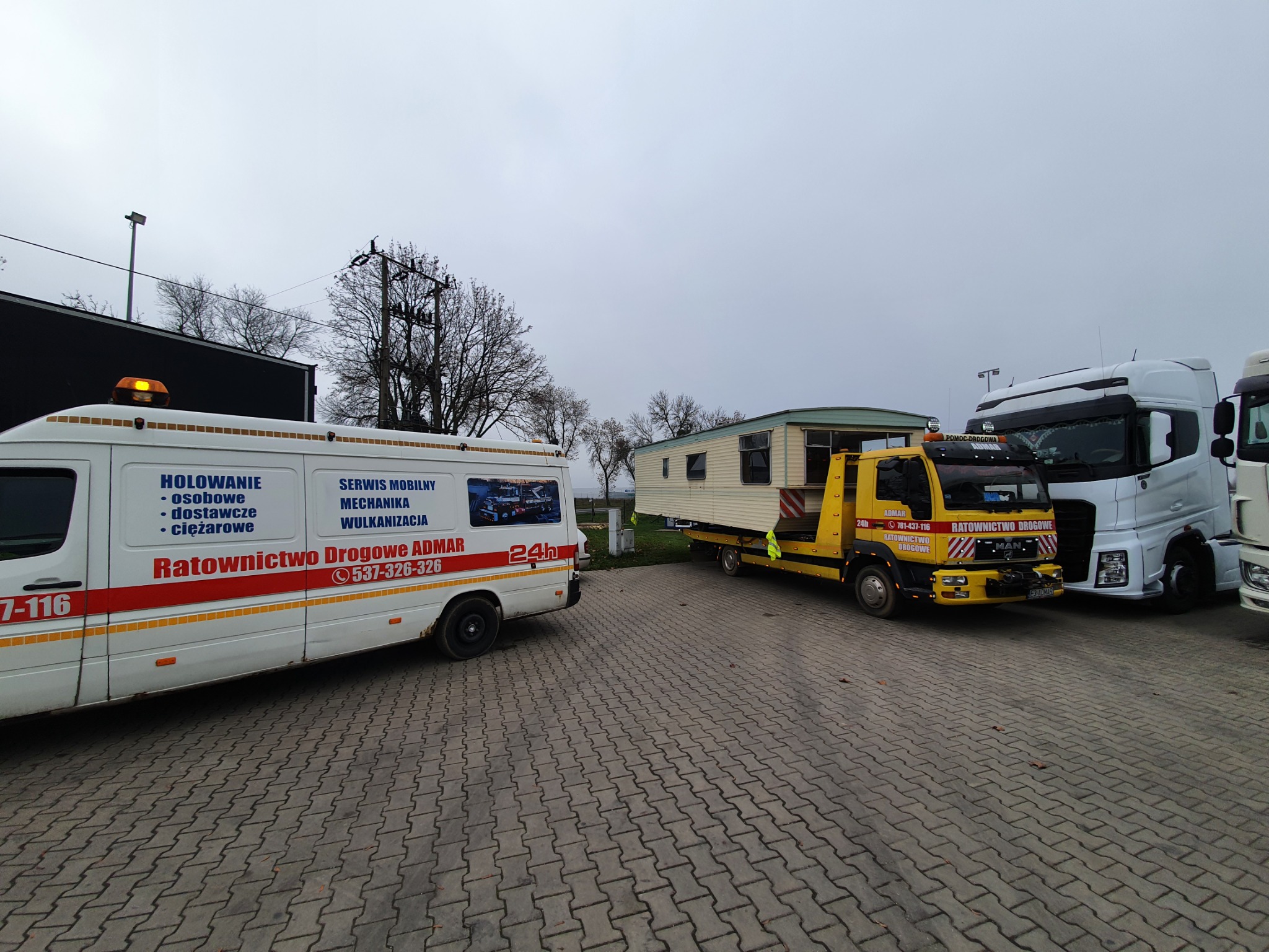Żółta laweta przewozi przyczepę kempingową na betonowym placu, obok zaparkowane białe busy i ciężarówki, w tle drzewa i pochmurne niebo.