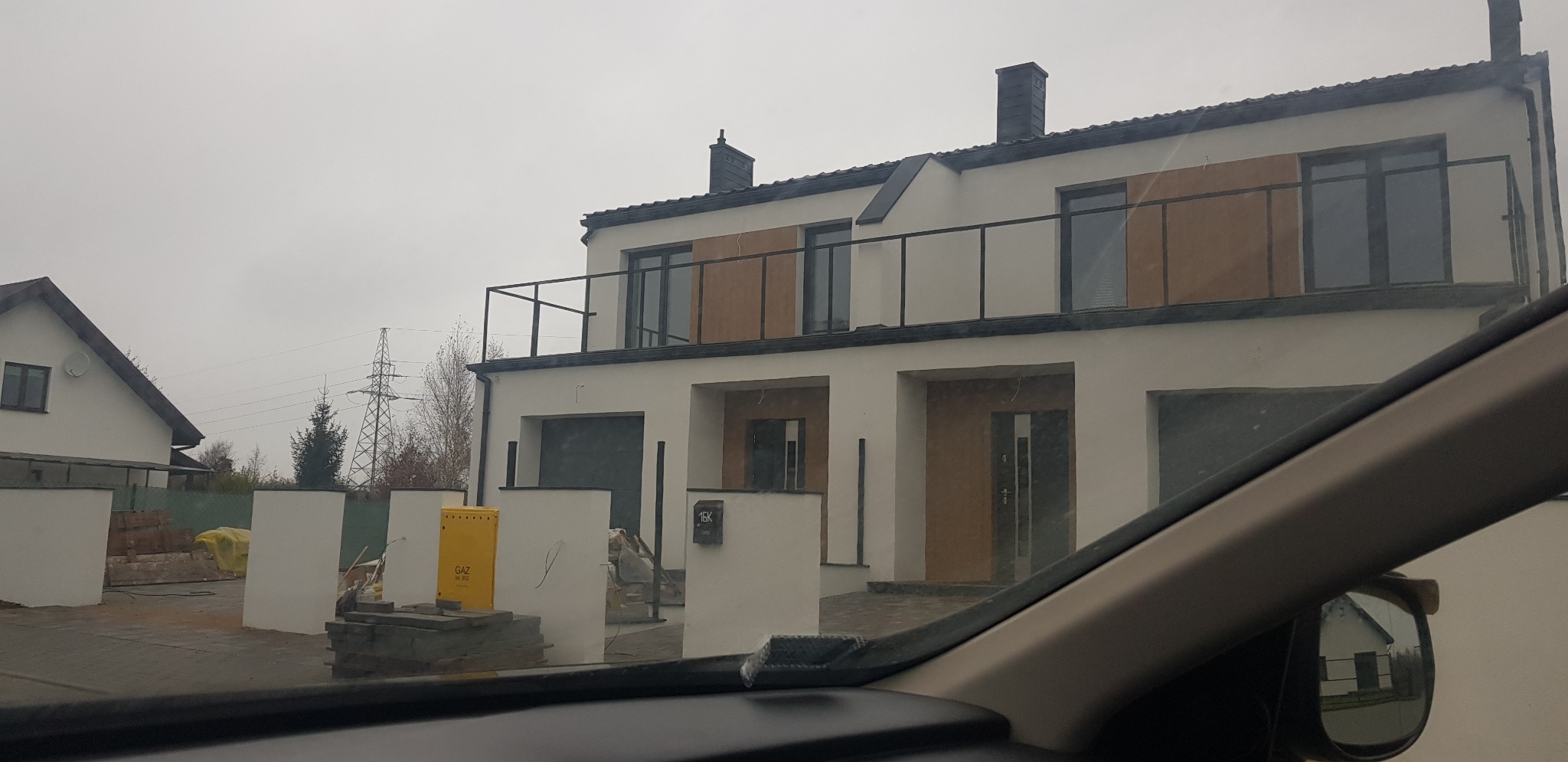 Dwukondygnacyjny dom bliźniak z balkonem i wykończoną elewacją, widok z wnętrza samochodu przez przednią szybę, widoczna skrzynka gazowa i materiały budowlane na posesji.