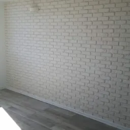 Ściana wykończona białymi płytkami imitującymi cegłę, z szarą podłogą z paneli drewnopodobnych i minimalistycznym oświetleniem sufitowym.