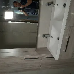 Nowoczesna łazienka z szarą armaturą, umywalką nablatową i dużym lustrem, w którym odbija się instalator robiący zdjęcie telefonem.