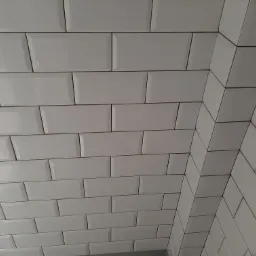 Ściana wyłożona błyszczącymi, białymi, prostokątnymi płytkami typu metro ułożonymi w przesuniętej konfiguracji, widok na narożnik, fuga w kolorze zbliżonym do płytek.